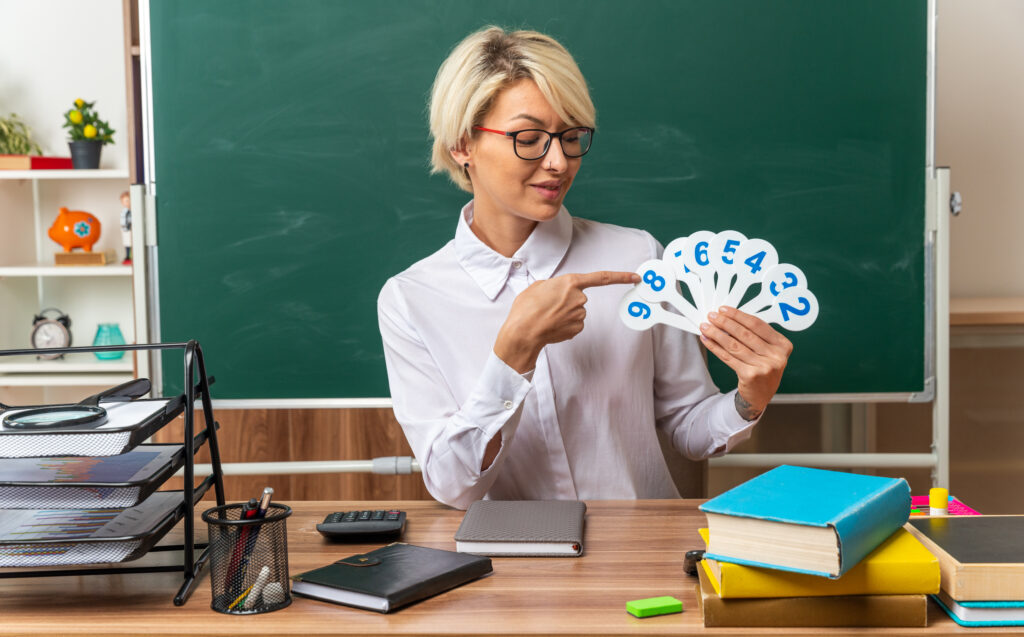 pleased young blonde female teacher wearing glasses sitting at desk with school tools in classroom showing and pointing at number fans looking at them